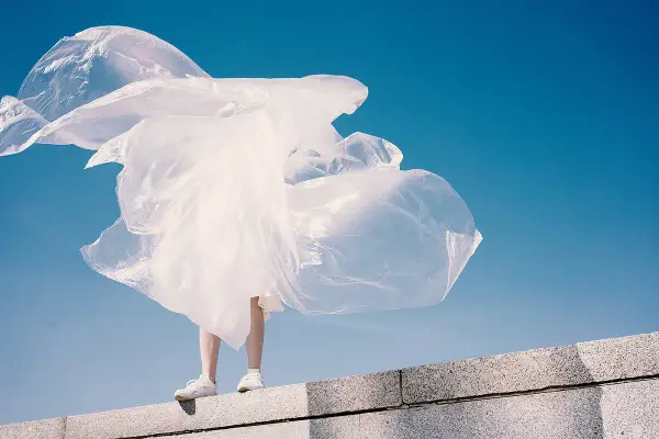 Eine Person steht auf einer Mauer, umgeben von wehendem, weißem Material, das an Plastik erinnert. Polyester schädlich für die Umwelt.