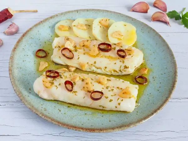 Dos filetes de merluza a la bilbaina, con patatas, ajo laminado y guindilla, en un plato azul.
