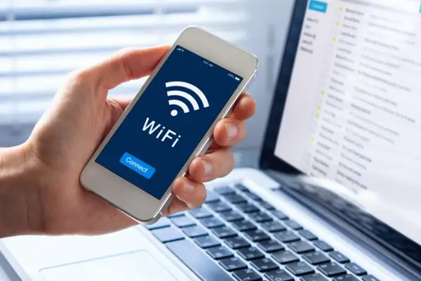 Dłoń trzyma smartfon z ikoną WiFi i napisem "Connect". Obok widać laptopa z listą e-maili, sugerującą pracę zdalną i dostęp do sieci, być może przez **ssid** domowy.