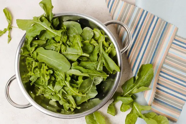 Rukola w durszlaku, idealna do przygotowania jadłospisu tygodniowego dieta FODMAP. Świeże liście na tle kolorowych tkanin.