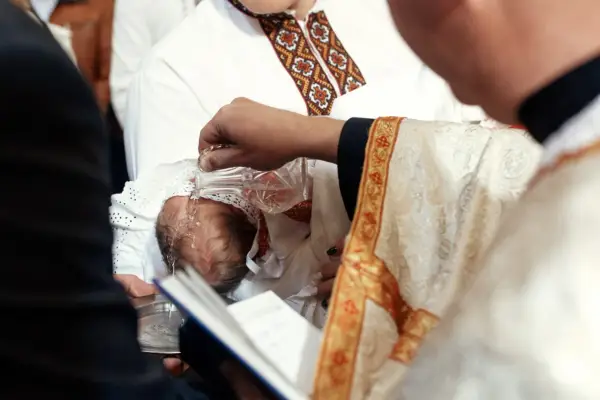 Chrzestni słuchają słów kapłana, gdy ten polewa wodą głowę niemowlęcia.