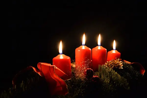 Fyra röda adventsljus brinner i mörkret, en del av en julkrans med granris och röda band. Detta är en visuell representation av vad betyder advent.
