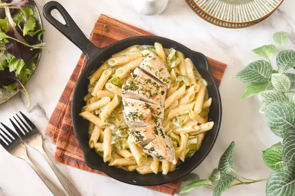 Deliciosa pasta con pollo en sartén, cubierta con hierbas frescas. Acompañada de una ensalada verde y tenedores.