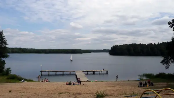 Spokojny dzień na plaży nad jeziorem, żaglówka na wodzie, drewniane molo. Pojezierze Brodnickie zaprasza do wypoczynku.
