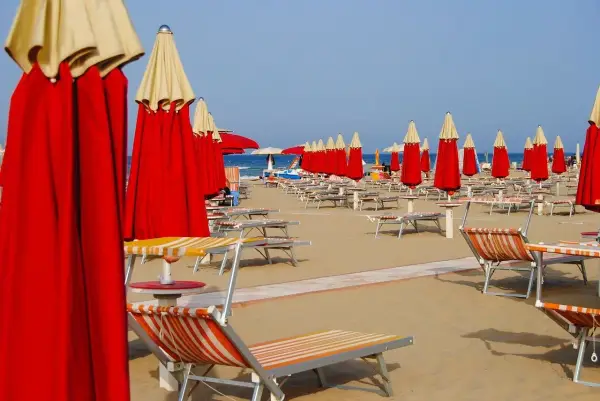 File di ombrelloni rossi e lettini prendisole su una spiaggia, forse a Riccione o Rimini, in attesa dei bagnanti.