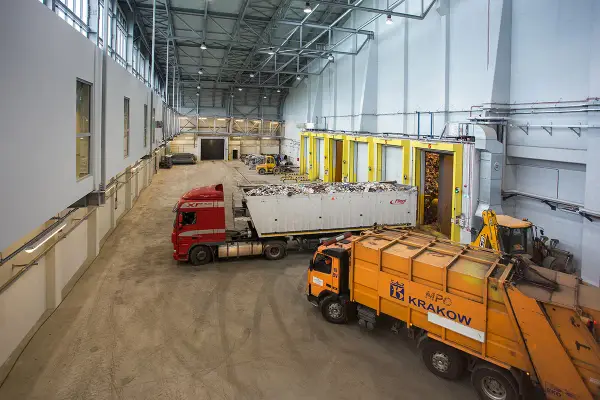 Wielka hala przemysłowa, gdzie trwa termiczne przekształcanie odpadów. Ciężarówki z odpadami czekają na rozładunek, a koparka przygotowuje materiał do przetworzenia.