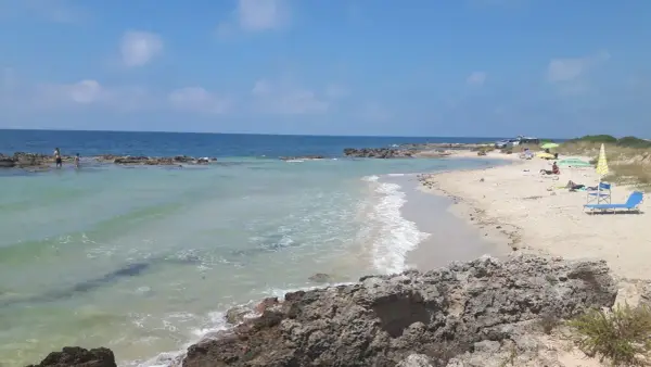 Spiaggia con scogli e mare cristallino, tipica delle calette puglia. Persone in acqua e sulla sabbia, ombrelloni e lettini.