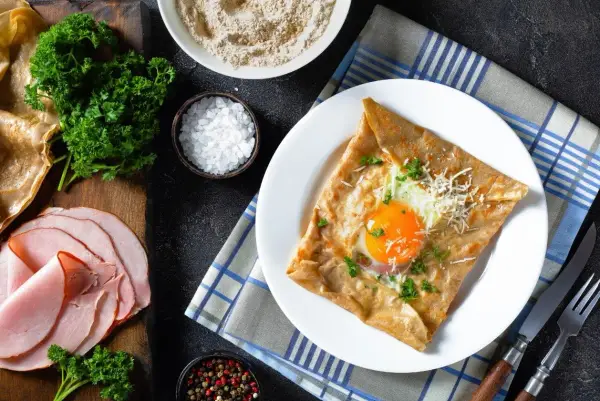 Francuska crepe z jajkiem sadzonym, serem i natką pietruszki na talerzu, obok szynka i składniki.