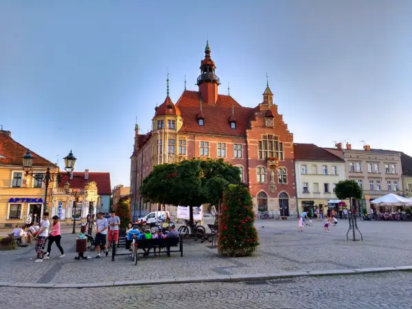 Rynek w Tarnowskich Górach tętni życiem. Ludzie spacerują, siedzą na ławkach, a dzieci bawią się na bruku. W tle widać zabytkowy ratusz.