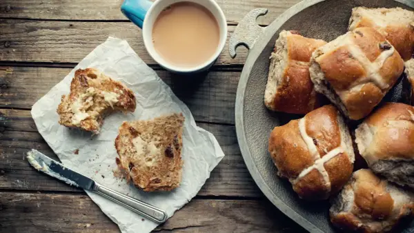 Na drewnianym stole leżą świeże, pachnące hot cross buns, obok kubek kawy i kromka posmarowana masłem.