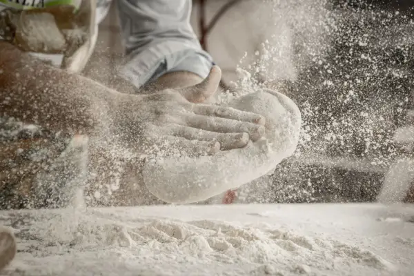 Ręce formują ciasto, a wokół unosi się chmura mąki na pizzę.