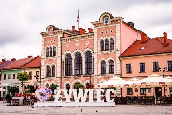 Duże białe litery "ŻYWIEC" na rynku, obok serca z ptaszkiem. W tle różowy ratusz i kawiarniane parasole.