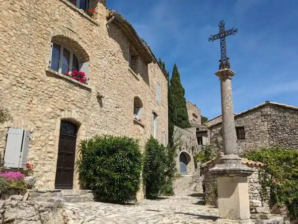 Village de pierre au soleil, le Crestet Vaucluse. Une croix orne la place pavée, entourée de maisons fleuries.