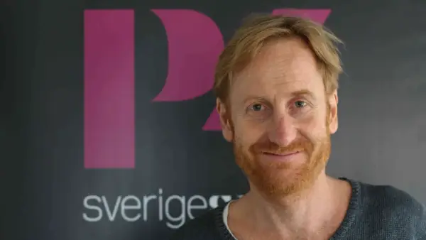 Gustaf Hammarsten, känd från filmer och tv-program, ler mot kameran framför en bakgrund med logotypen för Sveriges Television.
