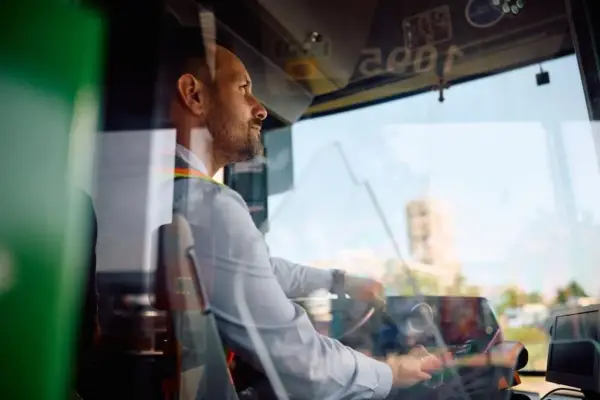 Skupiony kierowca autobusu prowadzi pojazd, trzymając kierownicę. Widać jego profil i fragment deski rozdzielczej.