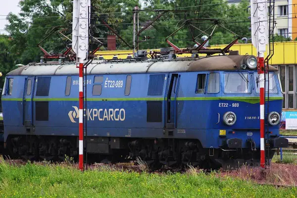 Niebieska lokomotywa PKP Cargo ET22-989 na tle zieleni i budynków.