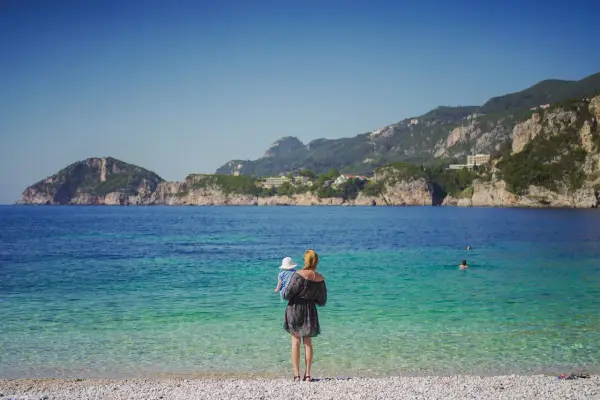 Kobieta z dzieckiem na rękach podziwia piękne Korfu plaże. Woda jest krystalicznie czysta, a w oddali widać góry.