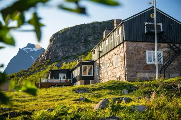 STF Kebnekaise fjällstation med stenfasad och mörk träpanel, belägen vid foten av ett berg. Grönskande sluttning i förgrunden.