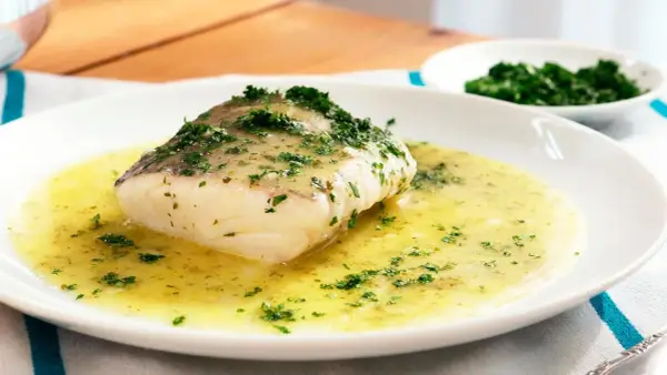 Un filete de merluza en salsa verde, con perejil picado, servido en un plato blanco.