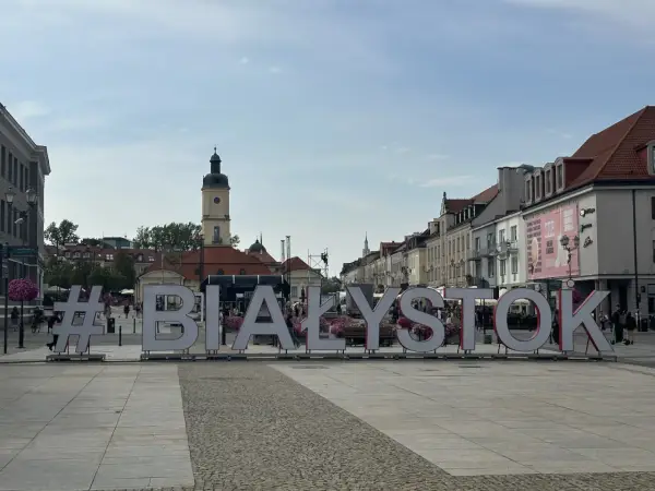 #Białystok na Rynku Kościuszki. Wielkie litery tworzą napis, w tle kamienice i wieża ratuszowa.