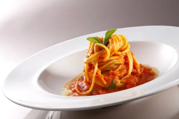 Un nido di spaghetti conditi con una salsa rossa vibrante di pomodorini freschi, guarnito con foglie di basilico verde brillante, servito in un piatto bianco elegante.