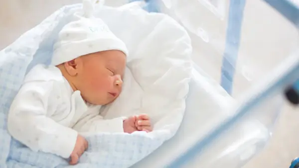 Śpiący noworodek w szpitalnym łóżeczku. Do wniosku o becikowe potrzebne są dokumenty potwierdzające urodzenie dziecka.