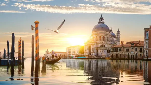 Góndola navega por canal veneciano al atardecer, con la Basílica de Santa Maria della Salute y otras **ciudades de Europa** reflejadas en el agua.