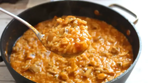 Cuchara sirviendo arroz caldoso con pollo y champiñones, humeante y apetitoso, de una sartén.