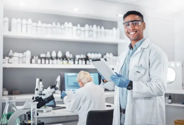 Uśmiechnięty technik farmacji w okularach ochronnych trzyma tablet w laboratorium, gdzie na półkach stoją liczne butelki, a w tle pracuje inna osoba.