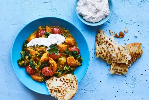 En blå skål med en mustig daal med sötpotatis, spenat och tomater, toppad med gräddfil. Serveras med naanbröd.