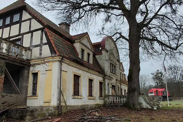 Zniszczony dworek w Paraszyne, z charakterystyczną architekturą i opadającą fasadą, otoczony drzewami.