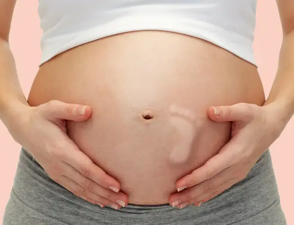 Brzuszek w 30 tygodniu ciąży, z widocznym odciskiem stópki dziecka. Ręce mamy delikatnie obejmują rosnący brzuch.