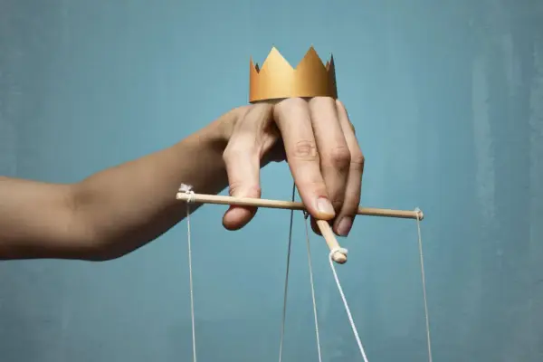 Dłoń z papierową koroną na palcach, sterująca marionetką. Symbol władzy i despoty.