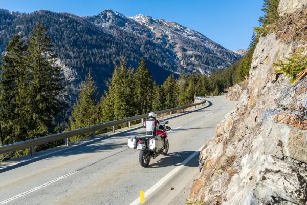 Motociclista su strada alpina in Svizzera, con montagne innevate e foreste. Ottimi consigli per un viaggio in moto in Svizzera.
