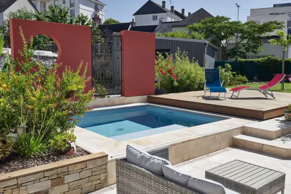 Une piscine moderne avec une belle décoration autour piscine : mur rouge, terrasse en bois, mobilier de jardin et végétation luxuriante.