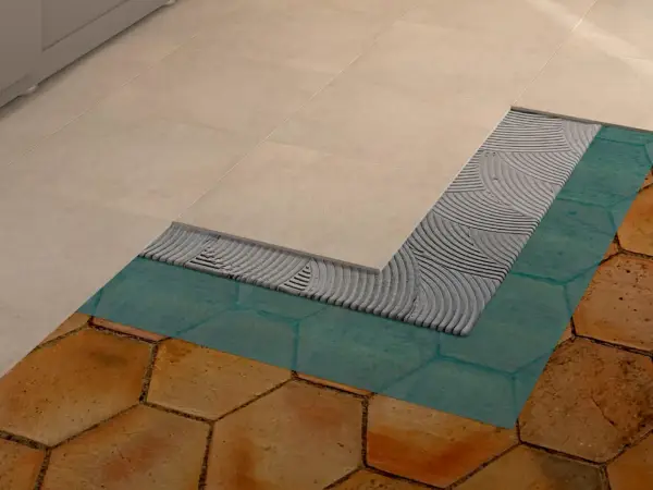 Dettaglio di un pavimento in fase di posa: nuove piastrelle beige sopra uno strato di colla, membrana impermeabilizzante turchese e vecchie piastrelle esagonali color terra.