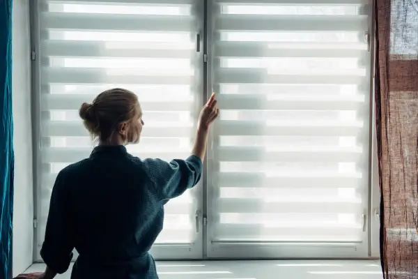 Une femme ajuste un store enrouleur blanc, laissant entrer la lumière du jour.