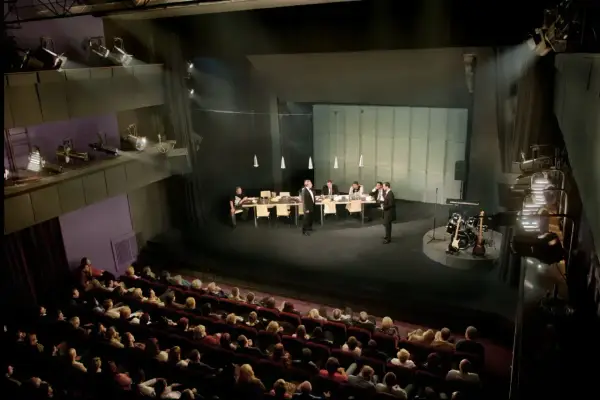 Scena teatralna z ludźmi przy stole i instrumentami muzycznymi. Widownia siedzi w ciemności, gotowa na spektakl. Dostawka w teatrze.