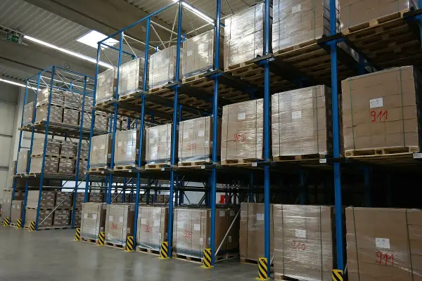 Wysoki magazyn centralny z regałami pełnymi paczek na paletach. Widać oznaczenia na kartonach, np. "911".