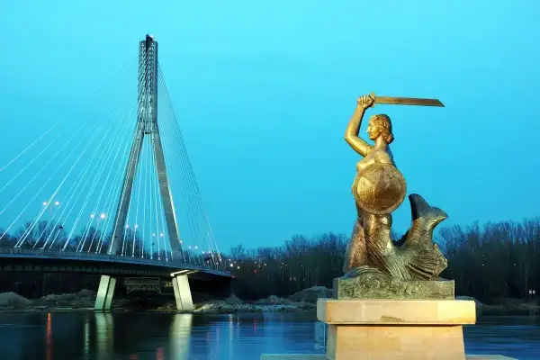 Syrenka warszawska z mieczem i tarczą stoi nad Wisłą, w tle nowoczesny most.