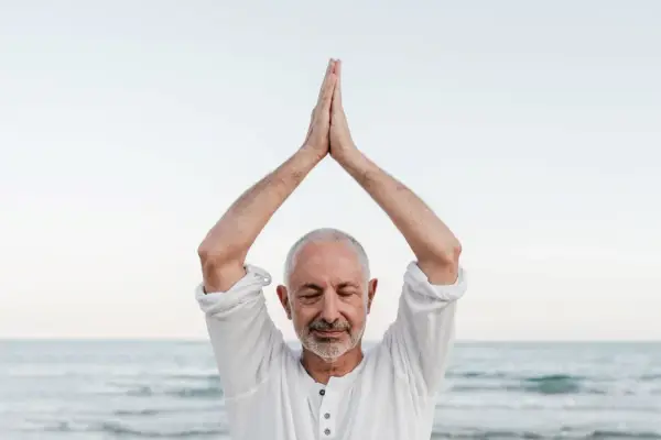Mężczyzna z zamkniętymi oczami, ręce złożone nad głową, praktykuje mindfulness co to. Spokój nad morzem.