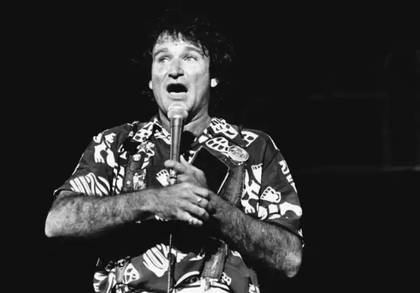 Robin Williams, w hawajskiej koszuli, śpiewa do mikrofonu, trzymając w dłoni notes.