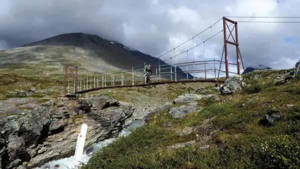 Hängebrucke über einem Fluss im schwedischen Gebirge. Ein Wanderer überquert die Brücke, umgeben von felsiger Landschaft und grüner Vegetation.