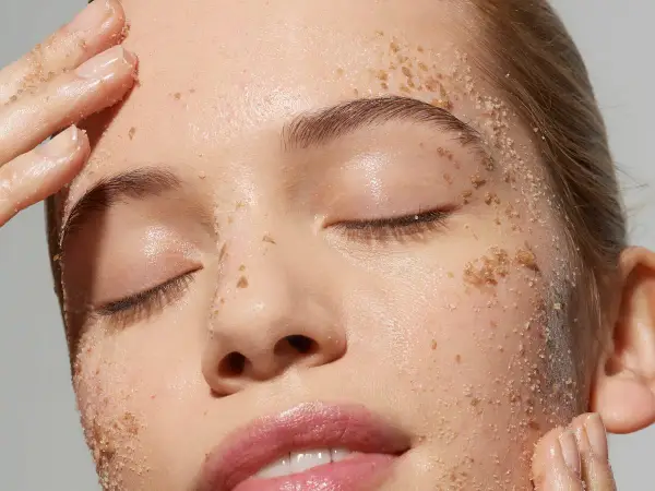 Una cara llena de granos de exfoliante, con dedos aplicándolo suavemente.