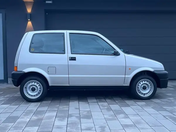 Srebrny Fiat Cinquecento na tle ciemnego garażu, z charakterystycznymi felgami i czarnymi zderzakami.
