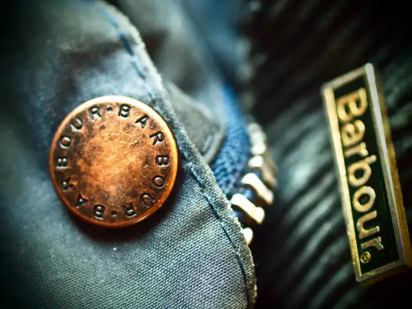 Detale kurtki Barbour: mosiężny guzik z logo i naszywka z nazwą firmy. Barbour to znana marka odzieży.