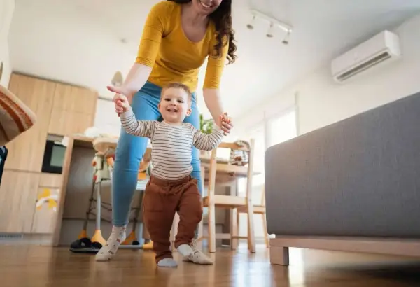Mamá ayuda a su bebé a dar sus primeros pasos en una habitación cómoda, con el aire acondicionado bebes funcionando para mantener la temperatura ideal.