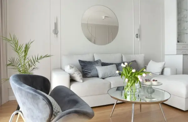 Przytulny salon z sofą i fotelami na tle jasnych, **białe ściany** zdobionych sztukaterią. Okrągłe lustro i bukiet tulipanów dodają elegancji.