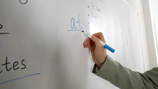 Mano scrive "at" su lavagna, spiegando il suo significato in inglese.