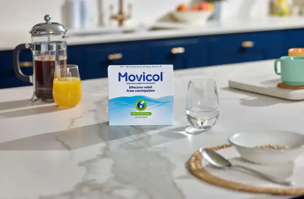 En låda Movicol med citron- och limsmak står på en köksbänk. Bredvid finns ett glas vatten, juice och en presskanna kaffe. Tänk på moxalole verkningstid för effektiv lindring.